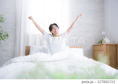 朝日を浴びるベッドで寝起きの若い男性　腕を伸ばしストレッチをする　　バンザイのポーズ 127882153