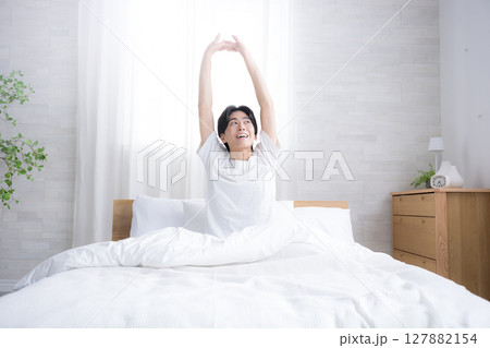 朝日を浴びるベッドで寝起きの若い男性 腕を伸ばしストレッチをする 朝日を浴びるベッドで寝起きの若い男性 腕を伸ばしストレッチをする 127882154