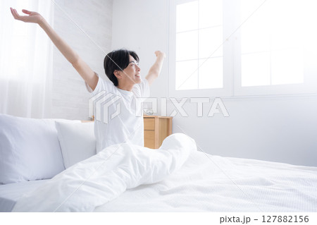 朝日を浴びるベッドで寝起きの若い男性　腕を伸ばしストレッチをする 見上げるバンザイのポーズ 127882156