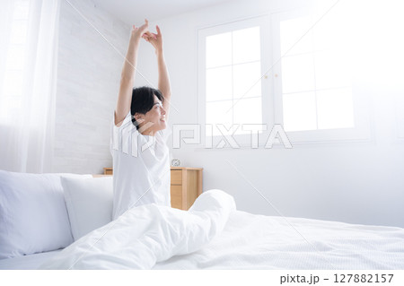 朝日を浴びるベッドで寝起きの若い男性　腕を伸ばしストレッチをする 127882157