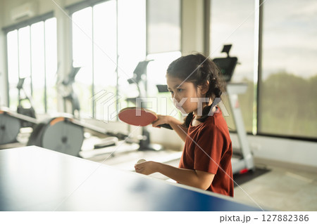 Young girl practicing table tennis at a modern gym setting with fitness equipment in the background and natural light streaming through large windows 127882386