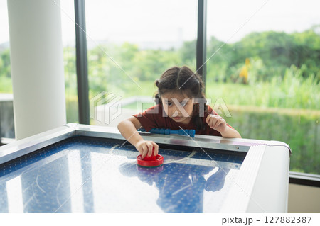 Young Girl Engaged in Air Hockey Game with Focus and Determination in Modern Leisure Space Surrounded by Lush Greenery Outdoors 127882387