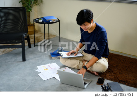 Man managing personal finances at home, reviewing reports with laptop and documents on the floor 127882451