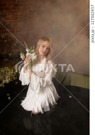 Young woman in white dress sitting in artificial pond with flowers and fog against brick wall Young woman in white dress sitting in artificial pond with flowers and fog against brick wall 127882657