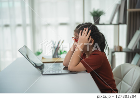 Frustrated young girl with glasses sitting at a desk, experiencing stress while working on a laptop in a bright, modern home office space 127883284