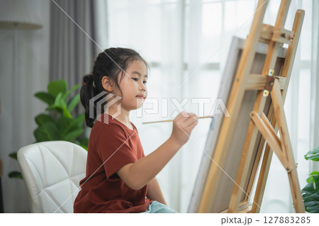 Young Girl Focused on Painting Creativity at Home with a Wooden Easel and Lush Green Plants in a Bright and Modern Interior Setting Young Girl Focused on Painting Creativity at Home with a Wooden Easel and Lush Green Plants in a Bright and Modern Interior Setting 127883285