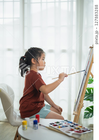 Young girl creating art at home, focused on painting with watercolors, sitting at a table by a sunny window, creative expression, childhood joy Young girl creating art at home, focused on painting with watercolors, sitting at a table by a sunny window, creative expression, childhood joy 127883288