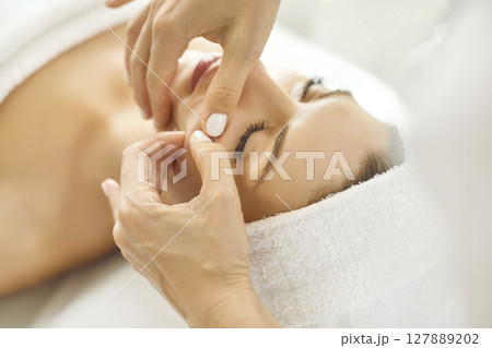 Female masseur making face relaxing massage to a young girl pinching the cheeks in beauty salon. Female masseur making face relaxing massage to a young girl pinching the cheeks in beauty salon. 127889202