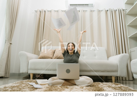 Asian woman throws papers in the living room as she finishes her work with her laptop on her lap. 127889393
