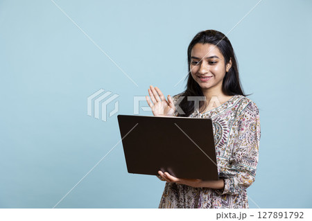 Confident upbeat girl sharing a story with best friend over web video call, standing against blue background with smartphone. Happy woman talking with mate online on teleconference. 127891792
