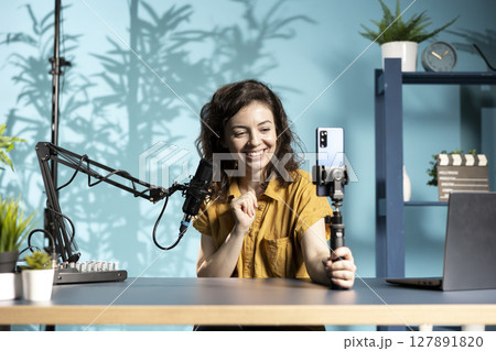 Cheerful internet star talking in front on cellphone attached to selfie stick in home studio, chatting with viewers. Happy woman using phone camera to record vlog for podcasting channel 127891820