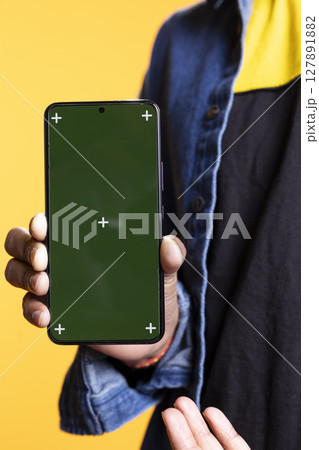 Indian content creator presenting green screen on mobile device, standing against orange background in the studio. Young male model wearing casual clothes and a man bun, internet channel. Indian content creator presenting green screen on mobile device, standing against orange background in the studio. Young male model wearing casual clothes and a man bun, internet channel. 127891882