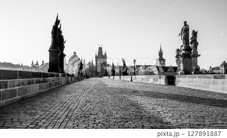 Sunrise casts a golden hue over Charles Bridge in Prague, revealing the cobblestone path and statues lining the bridge. The calm Vltava River reflects the city historic architecture. 127891887
