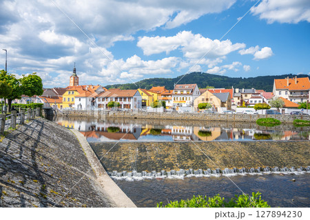 The tranquil waters of the Otava River reflect colorful buildings in Susice, Czechia. Visitors enjoy a sunny day with a backdrop of green hills and blue skies. 127894230