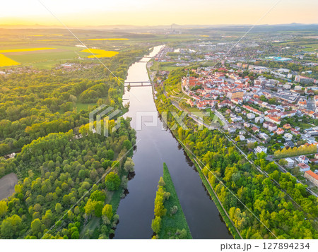 An aerial view captures the serene confluence of the Vltava and Labe rivers near Melnik. Lush greenery surrounds the winding rivers, leading to the picturesque town and vibrant fields at sunset. 127894234