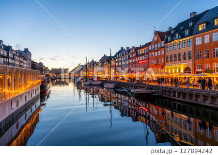 As dusk falls over Nyhavn, the waterfront glows with warm lights reflecting on the calm canal. People enjoy the vibrant atmosphere, dining and exploring the historic district. 127894242