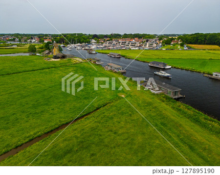 A picturesque aerial view of a Dutch windmill beside a canal with boats, surrounded by lush green fields, traditional houses, and distant waterways under a cloudy sky. A picturesque aerial view of a Dutch windmill beside a canal with boats, surrounded by lush green fields, traditional houses, and distant waterways under a cloudy sky. 127895190