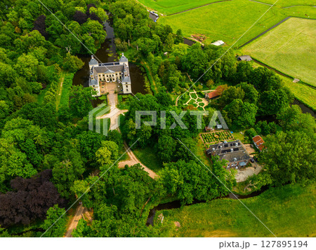 Aerial view of a historic castle surrounded by a forest, with vast green fields, winding waterways, and a river stretching across the flat Dutch countryside under a partly cloudy sky. 127895194