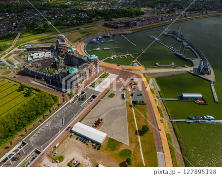 Aerial view of a Dutch harbor with modern residential buildings and green rooftops. Several docks and yachts line the calm water. The area features wide promenades, parking lots, and open spaces. Aerial view of a Dutch harbor with modern residential buildings and green rooftops. Several docks and yachts line the calm water. The area features wide promenades, parking lots, and open spaces. 127895198