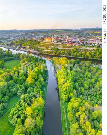 This aerial view captures Melnik City with its historic chateau perched above the Labe River. Lush greenery surrounds the riverbanks, showcasing the natural beauty of Czechia during sunset. 127896003