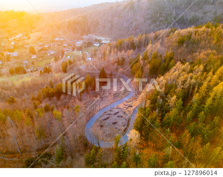 A winding road cuts through a forest of trees in autumn hues during sunset. The vibrant colors illuminate the landscape, creating a peaceful and serene atmosphere. A winding road cuts through a forest of trees in autumn hues during sunset. The vibrant colors illuminate the landscape, creating a peaceful and serene atmosphere. 127896014