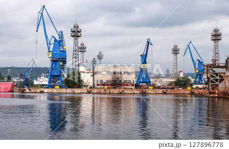 Industrial dockyard showcasing large blue cranes, buildings, and docked ships Industrial dockyard showcasing large blue cranes, buildings, and docked ships 127896778