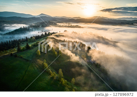 Aerial view of breathtaking sunrise over misty landscape with rolling hills and verdant fields. Soft light pierces through fog, casting long shadows and illuminating trees, creating magical scene. Aerial view of breathtaking sunrise over misty landscape with rolling hills and verdant fields. Soft light pierces through fog, casting long shadows and illuminating trees, creating magical scene. 127896972