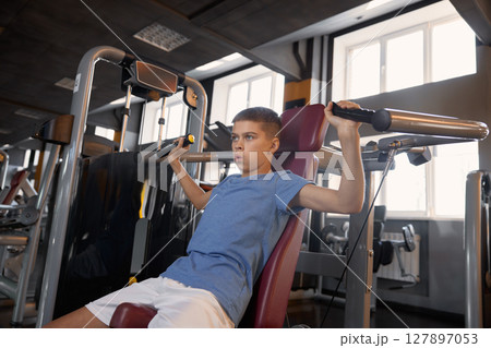 Teen boy doing sports exercise at machine for chest muscles pumping Teen boy doing sports exercise at machine for chest muscles pumping 127897053