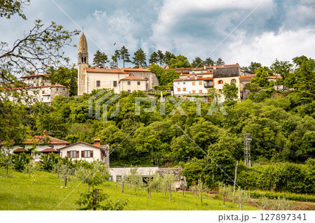 Scenic Hilltop Town Of Stanjel In Slovenia: Traditional Stone Houses And Historic Church Tower Rising Above Lush Greenery Under A Dramatic Cloudy Sky In The Karst Region 127897341