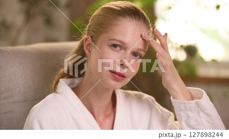 Woman in white robe touching forehead with relaxed expression indoors Woman in white robe touching forehead with relaxed expression indoors 127898244