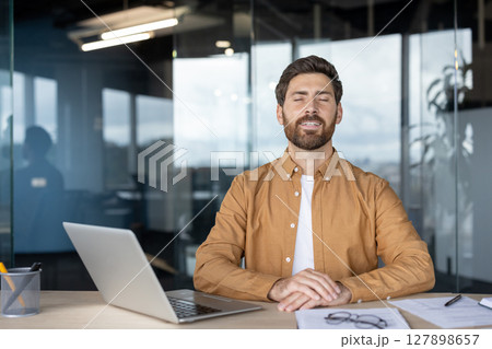 A man in an office setting with his eyes closed, looking relaxed, with a laptop and office supplies on the desk. A man in an office setting with his eyes closed, looking relaxed, with a laptop and office supplies on the desk. 127898657