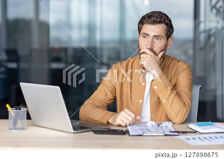 A thoughtful businessman ponders a problem in his office, with a laptop and documents nearby. A thoughtful businessman ponders a problem in his office, with a laptop and documents nearby. 127898675