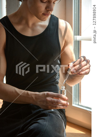 Man holding dropper bottle wearing black tank top by window in natural sunlight Man holding dropper bottle wearing black tank top by window in natural sunlight 127898914