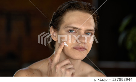 Young man applying skincare cream to face indoors with confident expression 127898944