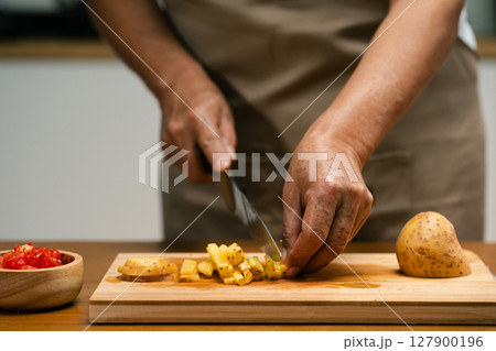 Close-up of Asian man's hands slicing potatoes on a wooden cutting board in the kitchen, preparing for cooking. Close-up of Asian man's hands slicing potatoes on a wooden cutting board in the kitchen, preparing for cooking. 127900196