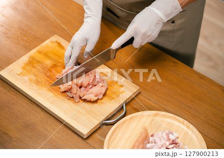 Close-up of Asian man's hands cutting chicken on a wooden cutting board in the kitchen, preparing for cooking. 127900213