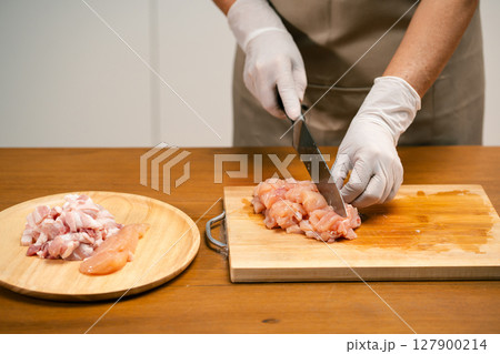 Close-up of Asian man's hands cutting chicken on a wooden cutting board in the kitchen, preparing for cooking. Close-up of Asian man's hands cutting chicken on a wooden cutting board in the kitchen, preparing for cooking. 127900214
