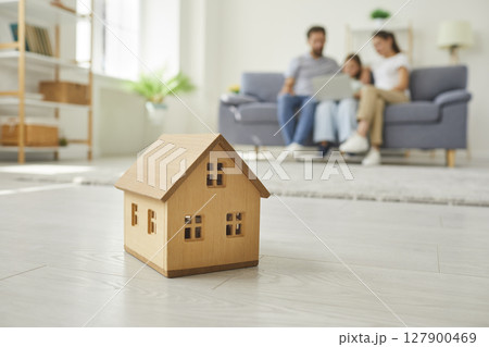 Wooden toy house on floor with a family of three sitting together on sofa in background. Wooden toy house on floor with a family of three sitting together on sofa in background. 127900469