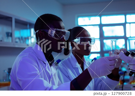 Two scientists wearing lab coats and safety glasses working together in a laboratory, carefully examining a test tube 127900728