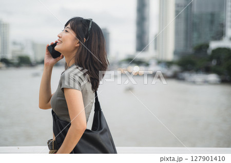 Happy solo Southeast Asian woman teenager travel blogger uses smartphone. An explorer enjoying local cultural experience Asia waiting for friend or express boat by Chao Phraya River Bangkok 127901410