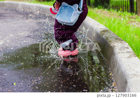 The child jumps through the puddles. The child in a bright cloak and colorful rubber boots joyfully jumps and splashes in a puddle next to the sidewalk, holding a small pink umbrella on a rainy day. The child jumps through the puddles. The child in a bright cloak and colorful rubber boots joyfully jumps and splashes in a puddle next to the sidewalk, holding a small pink umbrella on a rainy day. 127903166