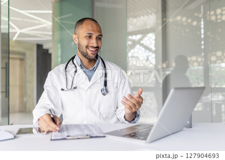 A doctor wearing a lab coat smiles while talking during a video conference from his office. He's using a laptop and gesturing. A doctor wearing a lab coat smiles while talking during a video conference from his office. He's using a laptop and gesturing. 127904693
