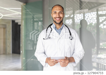 A smiling male doctor in a white coat, holding a tablet, standing in a modern healthcare setting. He looks friendly and approachable, ready to help. A smiling male doctor in a white coat, holding a tablet, standing in a modern healthcare setting. He looks friendly and approachable, ready to help. 127904733
