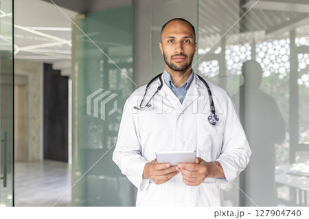 A doctor, wearing a stethoscope and lab coat, stands in an office holding a tablet. He appears calm and professional. 127904740