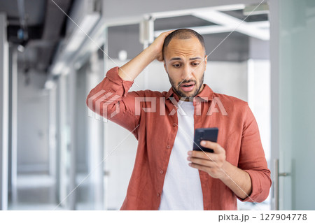 Sad disappointed man with phone in hands standing inside office. Businessman received fraudulent online notification message. Company employee with smartphone in hands reading frustrated. Sad disappointed man with phone in hands standing inside office. Businessman received fraudulent online notification message. Company employee with smartphone in hands reading frustrated. 127904778
