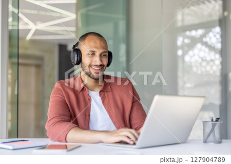 A smiling man with headphones on, working on a laptop in a modern office setting. Sunlight streams in from a nearby window. 127904789