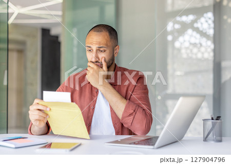 A concerned man is looking at a letter or document, his expression suggesting worry or concern. He is in an office setting with a laptop. A concerned man is looking at a letter or document, his expression suggesting worry or concern. He is in an office setting with a laptop. 127904796