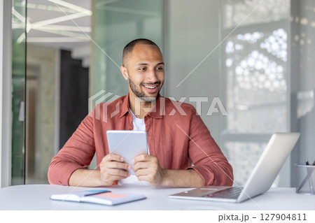 A smiling businessman sits at his desk, holding a tablet while working with a laptop in a modern office environment. Focus is on his expression. 127904811