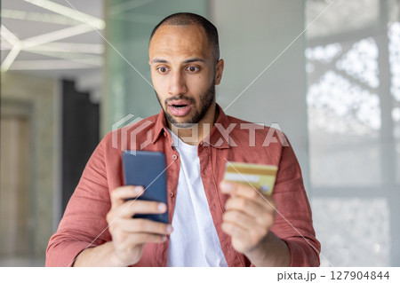 A surprised man looks at his phone while holding a credit card, expressing concern. He is likely reacting to financial news. 127904844