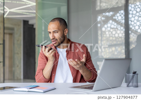 Worried businessman using voice assistant on his smartphone while sitting at his desk in a modern office setting with a laptop. 127904877